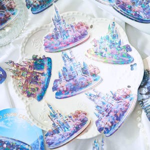Op de afbeelding: Een verzameling iriserende stickers met gedetailleerde illustraties van sprookjeskastelen. De stickers tonen een levendig kleurenpalet van blauw, paars en roze, met ingewikkelde architectonische details en omringende bomen. De stickers zijn gerangschikt op een wit, geschulpte bord.