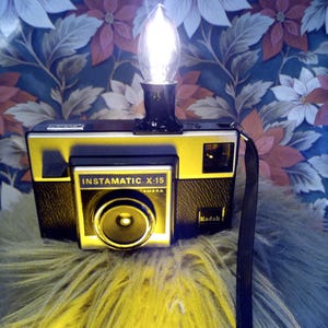 May include: A vintage Kodak Instamatic X-15 camera with a light bulb fixture on top. The camera is black and yellow with the text "Instamatic X-15 Camera" and the Kodak logo. The background features a floral pattern.