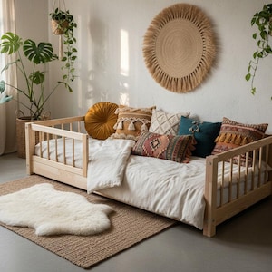 Op de afbeelding: Een licht houten daybed met een wit dekbed en decoratieve kussens in diverse kleuren en patronen. Een geweven vloerkleed en een schapenvachtkleed liggen op de vloer. Planten in hangmanden en potten geven de kamer een natuurlijke uitstraling.