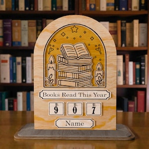 Puede incluir: Contador de libros de madera con una pila de libros, velas y detalles de estrellas. El contador muestra "Libros leídos este año" con los números 907. También incluye un espacio para un nombre.