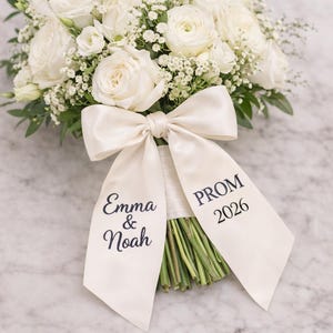 May include: A floral bouquet of white roses and other flowers, secured with a cream satin ribbon. The ribbon is printed with the names "Emma & Noah" and "PROM 2026" in dark blue script. The arrangement is set against a marble surface.