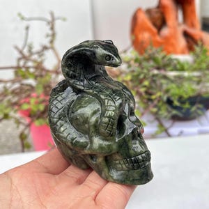 Op de afbeelding: Een donkergroene, gebeeldhouwde stenen sculptuur van een schedel met een cobra slang eromheen gewikkeld. De kop van de slang is omhoog en de schedel heeft gedetailleerde kenmerken. De sculptuur wordt in een hand gehouden.