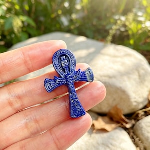 May include: A deep blue lapis lazuli stone carved into the shape of an Egyptian ankh symbol. The surface features intricate engraved details including a central winged figure and geometric patterns, showcasing the natural dark blue color.