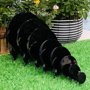 Może przedstawiać: Zestaw siedmiu czarnych akrylowych stojaków wystawowych o różnych rozmiarach, ułożonych w kolejności malejącej. Każdy okrągły stojak jest podparty trzema małymi nóżkami. Stojaki umieszczono na zielonej sztucznej trawie, z donicą w tle.