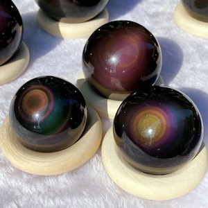 May include: A collection of iridescent obsidian spheres, displaying swirling patterns in purple, gold, and green hues. Each sphere rests on a small, round, light-coloured wooden stand. The spheres have a polished, reflective surface.