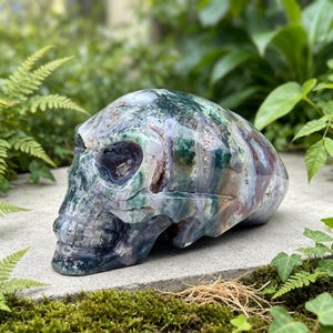 May include: A polished skull carving made from moss agate, featuring a complex pattern of deep forest green, translucent white, and earthy brown inclusions throughout the stone, with a smooth, glossy finish and defined anatomical features.