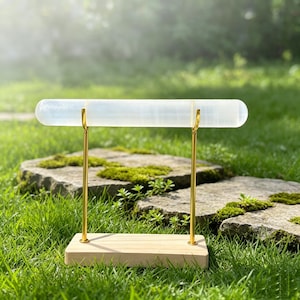 May include: A jewellery display stand featuring a horizontal, translucent white cylindrical bar supported by two thin, vertical gold-toned metal rods attached to a rectangular, light-coloured wooden base, designed for showcasing bracelets or watches.