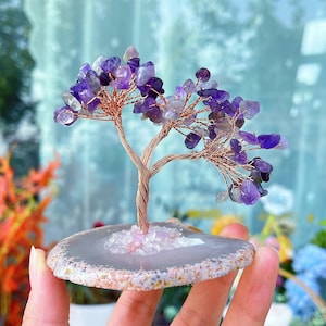 Könnte beinhalten: Eine handgefertigte Amethyst-Edelsteinbaum-Skulptur. Der Baum hat einen Stamm und Äste aus Kupferdraht, die mit Amethystchips verziert sind. Er ist auf einer grauen Achatscheibenbasis montiert. Die Amethyststeine ​​reichen von hell- bis tiefviolett.