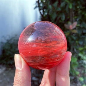 Könnte beinhalten: Eine klare, rote Kristallkugel mit inneren Streifen. Die Kugel wird in einer Hand gehalten, mit einem verschwommenen grünen Hintergrund aus Blättern. Der Kristall hat eine polierte, reflektierende Oberfläche, die die roten und braunen Muster zeigt.