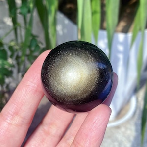 Può includere: Una gemma di ossidiana dorata lucidata e sferica. La pietra scura, quasi nera, ha un'iridescenza dorata scintillante sulla sua superficie. La sfera è tenuta in una mano, con uno sfondo verde sfocato.