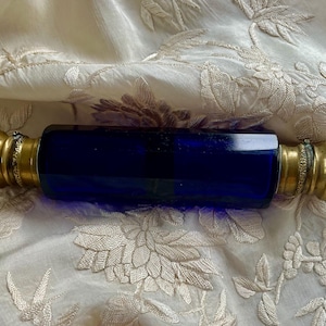 May include: A cobalt blue glass perfume bottle with ornate gold-colored metal ends. The bottle is rectangular with rounded edges. The metal ends have a floral design. The bottle is displayed on a cream-colored, embroidered fabric.