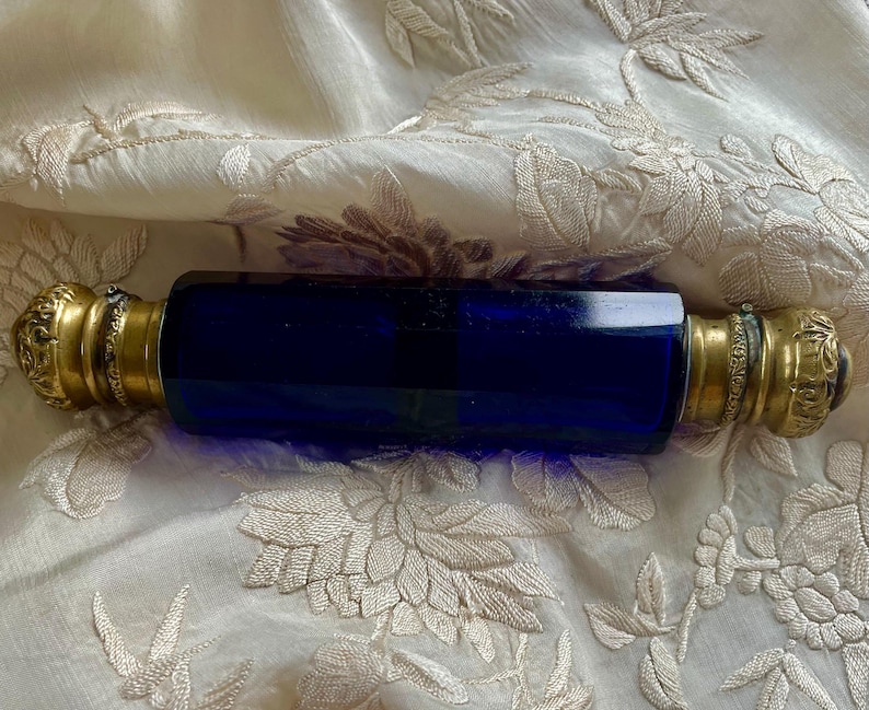 May include: A cobalt blue glass perfume bottle with ornate gold-colored metal ends. The bottle is rectangular with rounded edges. The metal ends have a floral design. The bottle is displayed on a cream-colored, embroidered fabric.