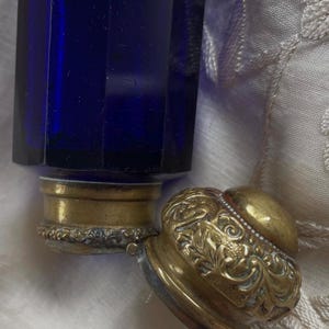May include: A cobalt blue glass perfume bottle with a brass base and a decorative, ornate brass lid. The lid is open, revealing the bottle's opening. The bottle has a faceted design.
