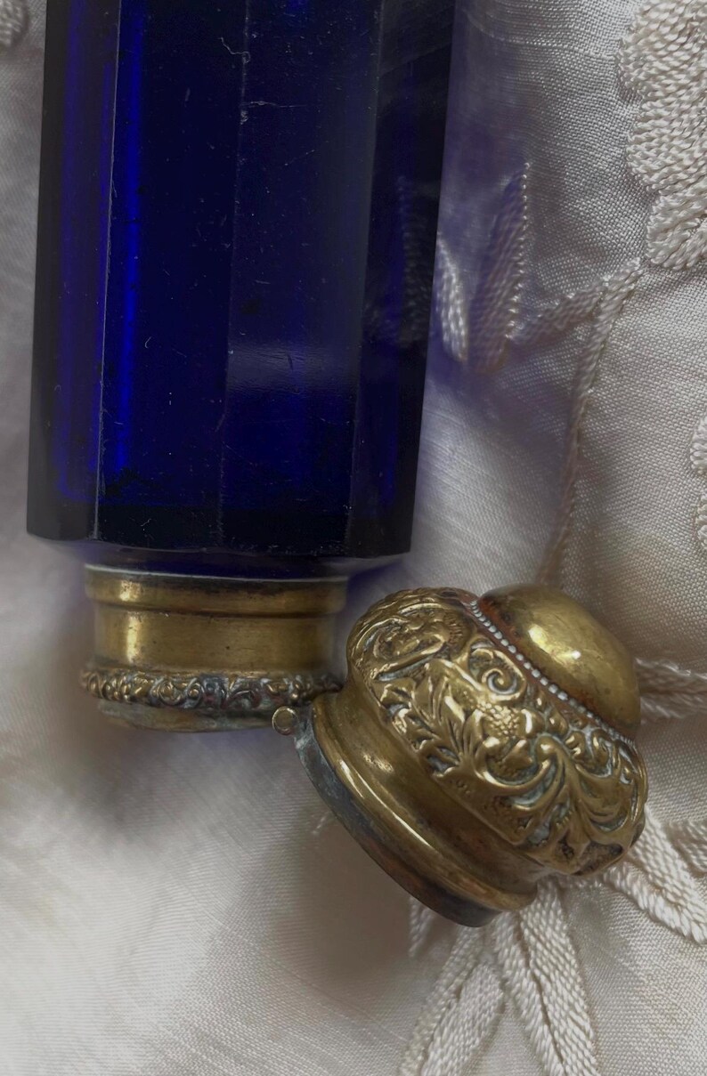 May include: A cobalt blue glass perfume bottle with a brass base and a decorative, ornate brass lid. The lid is open, revealing the bottle's opening. The bottle has a faceted design.
