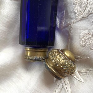May include: A cobalt blue glass perfume bottle with a gold-colored metal base and a decorative, ornate lid. The lid is detached, revealing the bottle's opening. The bottle has a geometric, faceted design.