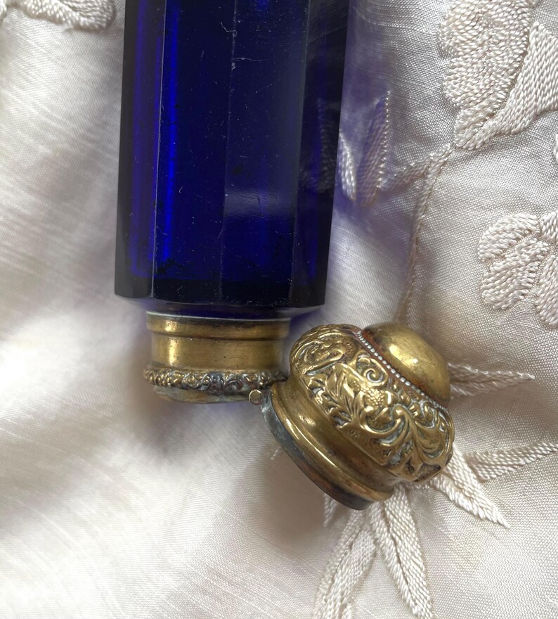 May include: A cobalt blue glass perfume bottle with a gold-colored metal base and a decorative, ornate lid. The lid is detached, revealing the bottle's opening. The bottle has a geometric, faceted design.