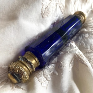 May include: A cobalt blue glass perfume bottle with a faceted body and ornate brass fittings. The bottle is lying on a cream-colored, embroidered fabric. The brass components include a decorative cap and a ring around the neck.