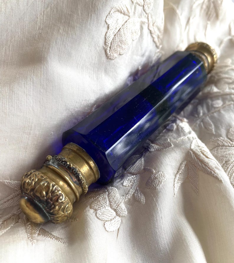May include: A cobalt blue glass perfume bottle with a faceted body and ornate brass fittings. The bottle is lying on a cream-colored, embroidered fabric. The brass components include a decorative cap and a ring around the neck.