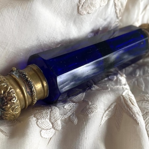 May include: A vintage perfume bottle with a faceted, deep blue glass body and ornate brass fittings. The bottle has a hexagonal shape and is topped with a decorative, floral-patterned cap. The brass has a warm, antique finish.