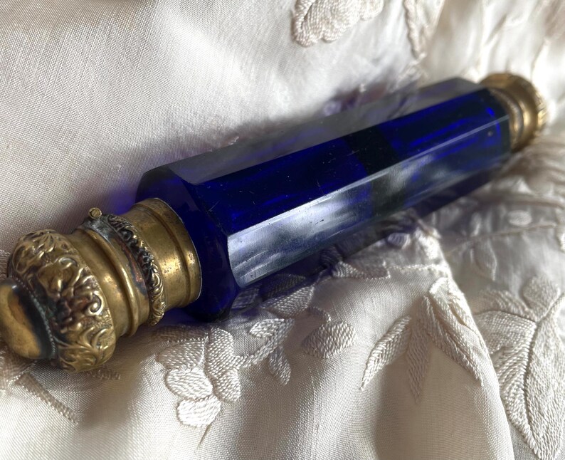 May include: A vintage perfume bottle with a faceted, deep blue glass body and ornate brass fittings. The bottle has a hexagonal shape and is topped with a decorative, floral-patterned cap. The brass has a warm, antique finish.