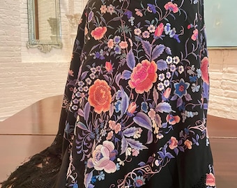 May include: A black shawl with a vibrant floral pattern in shades of pink, orange, blue, and purple. The shawl features a detailed design of flowers and leaves, with a fringed edge. The shawl is draped over a mannequin, showcasing its intricate embroidery.