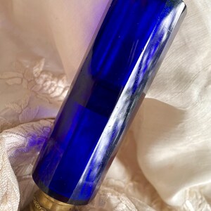 May include: A cobalt blue glass perfume bottle with a brass top and base. The bottle has a faceted, rectangular shape. The brass components have an ornate, antique design. The bottle is set against a cream-colored fabric.