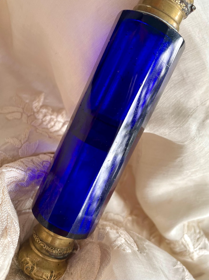 May include: A cobalt blue glass perfume bottle with a brass top and base. The bottle has a faceted, rectangular shape. The brass components have an ornate, antique design. The bottle is set against a cream-colored fabric.