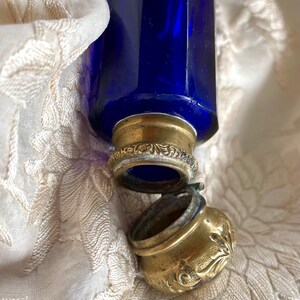 May include: A cobalt blue glass bottle with a brass-colored metal cap and decorative accents. The bottle is partially open, revealing the interior. The bottle rests on a cream-colored, patterned fabric.