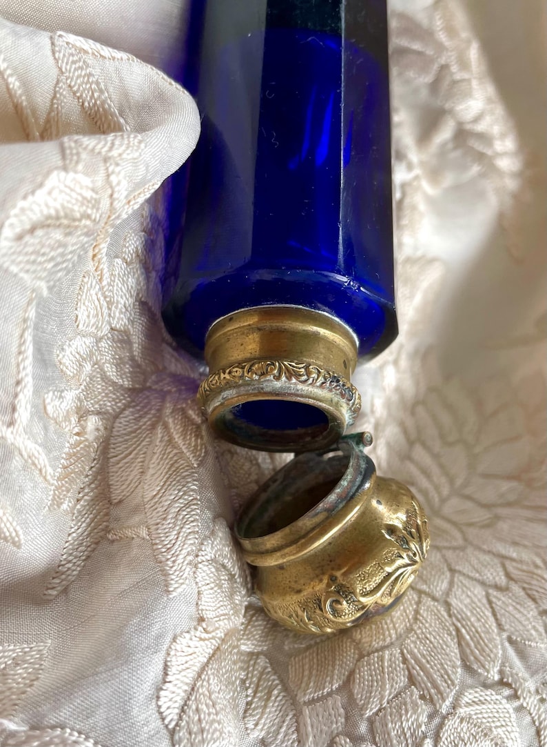 May include: A cobalt blue glass bottle with a brass-colored metal cap and decorative accents. The bottle is partially open, revealing the interior. The bottle rests on a cream-colored, patterned fabric.