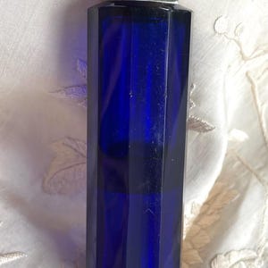 May include: A vintage cobalt blue perfume bottle with a faceted body and ornate gold-toned metal accents. The bottle has a decorative stopper and base. The bottle is likely made of glass and is displayed on a white, textured surface.