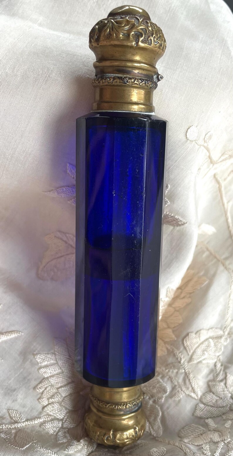May include: A vintage cobalt blue perfume bottle with a faceted body and ornate gold-toned metal accents. The bottle has a decorative stopper and base. The bottle is likely made of glass and is displayed on a white, textured surface.
