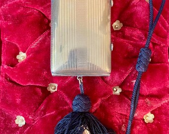 May include: A rectangular metallic cigarette case featuring a fine vertical striped texture. The case is attached to a dark blue braided cord that loops at the top and terminates at the bottom in a large, dense dark blue tassel with a metal tip.