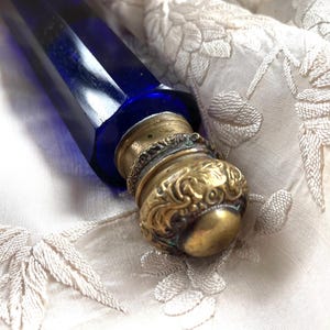 May include: A cobalt blue glass bottle with a brass stopper. The stopper has an ornate, embossed design. The bottle is lying on a cream-colored, embroidered fabric. The bottle's shape is long and angular.