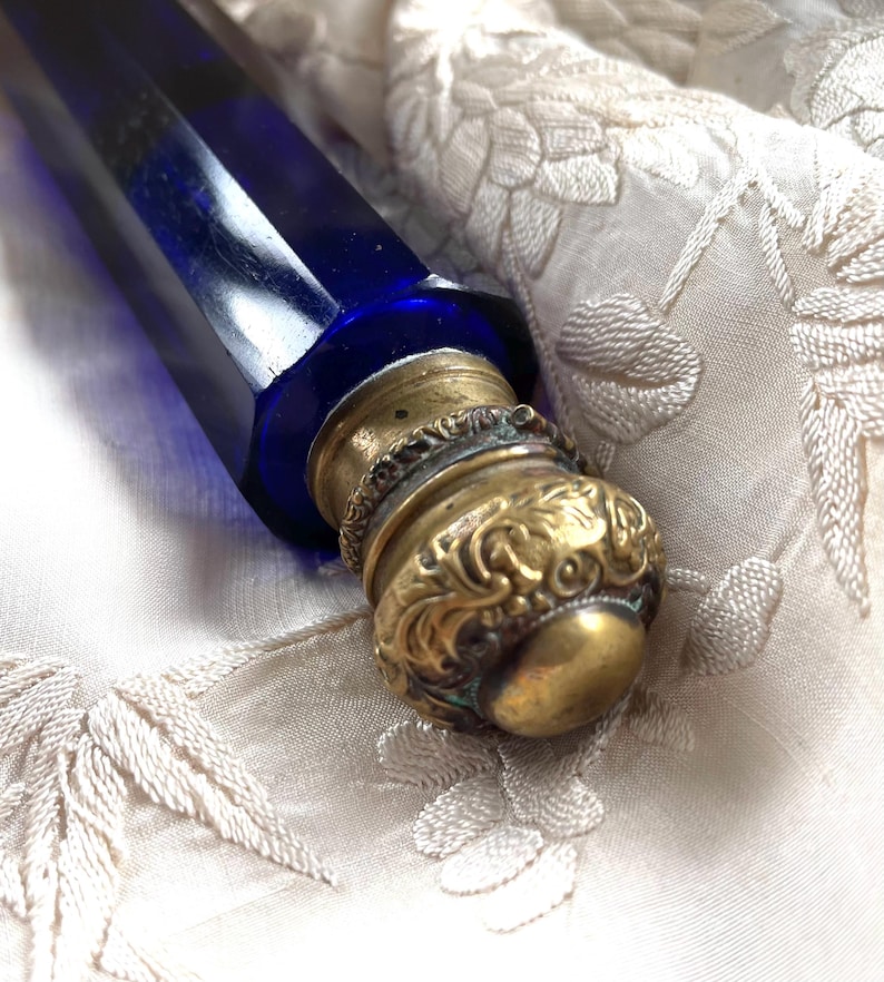 May include: A cobalt blue glass bottle with a brass stopper. The stopper has an ornate, embossed design. The bottle is lying on a cream-colored, embroidered fabric. The bottle's shape is long and angular.