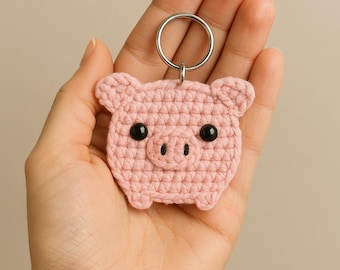 May include: A handmade pink crochet pig keychain with black safety eyes and a silver keyring. The pig has a simple, rounded design with small ears and a snout. The keychain is held in a hand against a neutral background.
