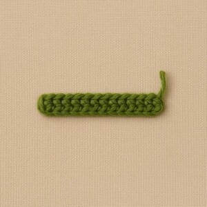 Op de afbeelding: Een close-up van een groene haaksteek. De steek heeft een rechthoekige vorm met een gebogen uiteinde en een enkele steek die omhoog steekt. De achtergrond is een neutrale beige stof.