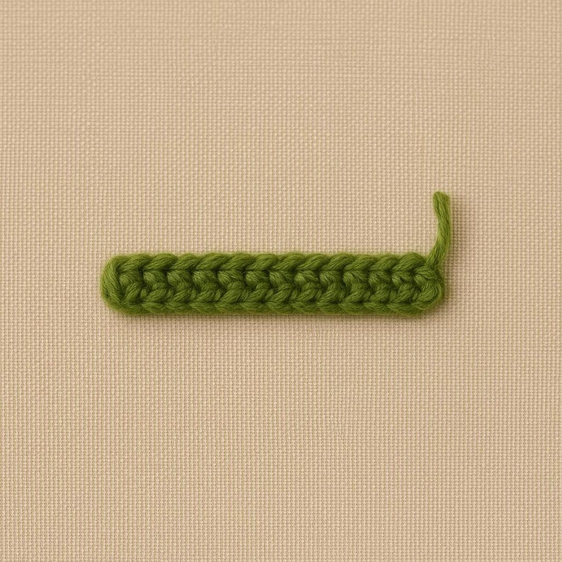 Op de afbeelding: Een close-up van een groene haaksteek. De steek heeft een rechthoekige vorm met een gebogen uiteinde en een enkele steek die omhoog steekt. De achtergrond is een neutrale beige stof.