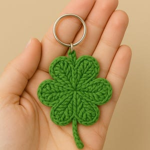 Op de afbeelding: Een groene, gehaakte klavervier sleutelhanger met een zilverkleurige ring. De klaver is gemaakt van getextureerd garen en wordt tegen een neutrale achtergrond gehouden.