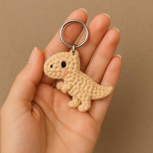 Op de afbeelding: Een gehaakte dinosaurus sleutelhanger in een lichtbruine kleur. De dinosaurus heeft zwarte ogen en roze wangen. De sleutelhanger heeft een zilveren metalen ring.
