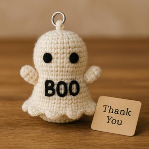 Puede incluir: Un llavero de fantasma tejido a ganchillo en blanco roto con ojos negros y la palabra "BOO" en negro. El llavero tiene un anillo plateado en la parte superior. Una peque&ntilde;a tarjeta marr&oacute;n con "Thank You" impresa est&aacute; al lado del fantasma.