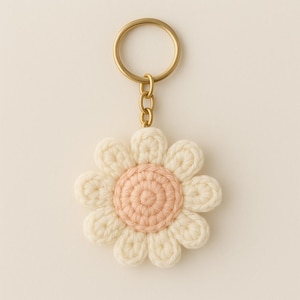 Könnte beinhalten: Ein handgefertigter Schlüsselanhänger mit einer gehäkelten Blume, einem Messingring und einer Kette. Die Blume hat eine cremefarbene Außenschicht und eine hellrosa Mitte. Ein bezauberndes Accessoire.