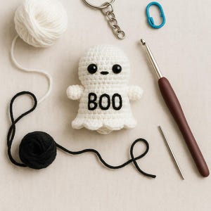 Puede incluir: Un llavero de fantasma de ganchillo blanco hecho a mano con la palabra "BOO" en negro. La imagen tambi&eacute;n incluye una madeja de hilo blanco, una madeja de hilo negro, un ganchillo, una aguja lanera y un marcador de punto azul.