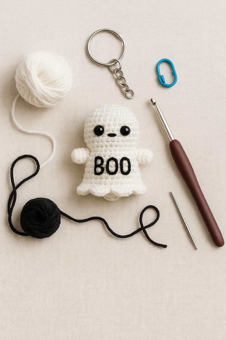 Puede incluir: Un llavero de fantasma de ganchillo blanco hecho a mano con la palabra "BOO" en negro. La imagen tambi&eacute;n incluye una madeja de hilo blanco, una madeja de hilo negro, un ganchillo, una aguja lanera y un marcador de punto azul.