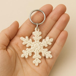 May include: A cream-coloured, crocheted snowflake keyring. The snowflake design features six symmetrical points, each with intricate detailing. The keyring has a silver-coloured metal ring for attaching to keys or bags. The snowflake is approximately 7.6 cm in diameter.