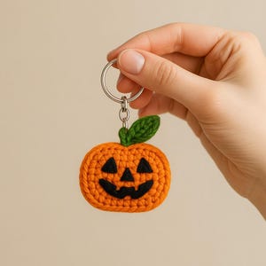 May include: A crocheted pumpkin keychain in orange with a black jack-o'-lantern face and a green leaf. The keychain is attached to a silver metal ring. A festive, handmade item perfect for Halloween.