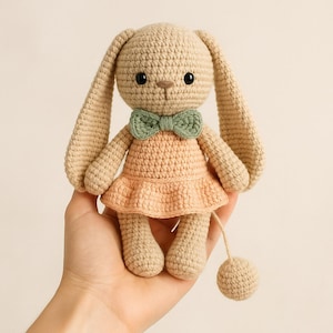 May include: A handmade crochet bunny doll in beige with a peach-colored dress and a green bow tie. The bunny has long ears, black eyes, and a small ball attached to a string. A charming, soft toy.