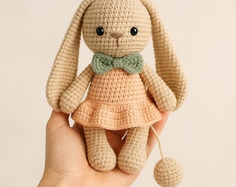 Puede incluir: Muñeca de conejo de ganchillo beige, con un vestido color melocotón y una pajarita verde. El conejo tiene orejas largas y ojos negros. Una pequeña bola está unida a una cuerda. Un juguete suave y hecho a mano.