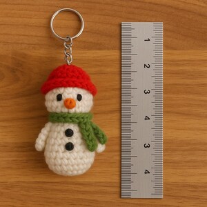 Op de afbeelding: Een handgemaakte gehaakte sneeuwpop sleutelhanger. De sneeuwpop heeft een rode hoed, groene sjaal en zwarte knoopogen en mond. De sneeuwpop is ongeveer 5 cm hoog. De sleutelhanger is bevestigd aan een zilveren metalen ring.