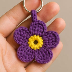 Może przedstawiać: Ręcznie robiony brelok do kluczy w kształcie kwiatka zrobionego na szydełku, w kolorze fioletowym i żółtym. Kwiat ma pięć płatków i żółty środek. Srebrny metalowy pierścień jest przymocowany do kluczy. Ozdobny dodatek.