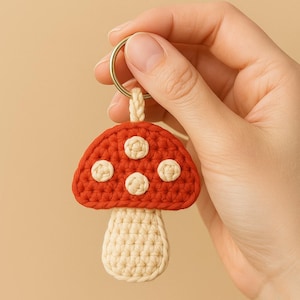 May include: A handmade crochet mushroom keychain. The mushroom cap is red with three white spots, and the stem is beige. The keychain is attached to a gold-colored ring.